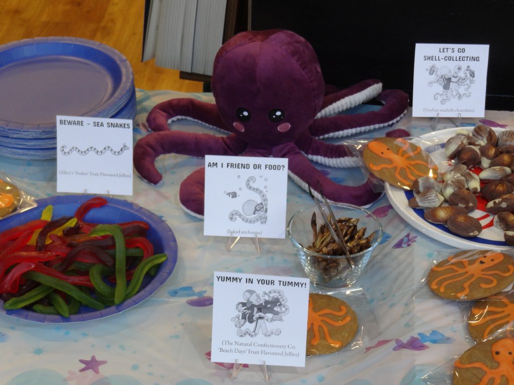 Refreshments table with purple octopus guarding jelly snakes, gingerbread, anchovies and seashell chocolates. Little signs use illustrations from the book.